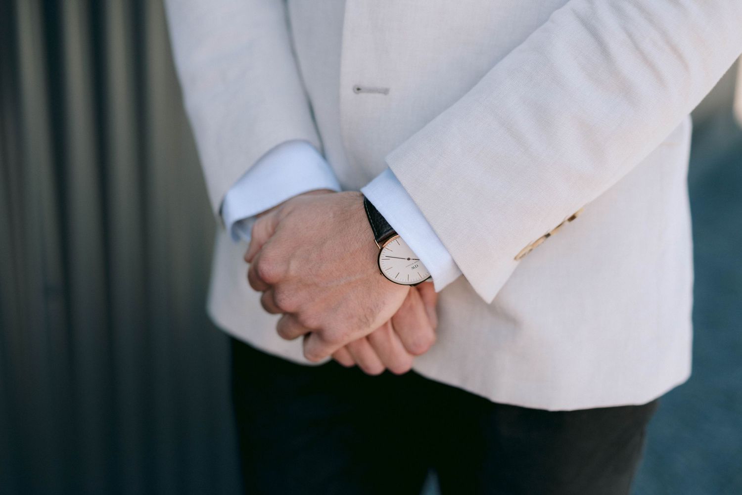 Close up of someone wearing a white suit jacket with clasped hands behind their back.