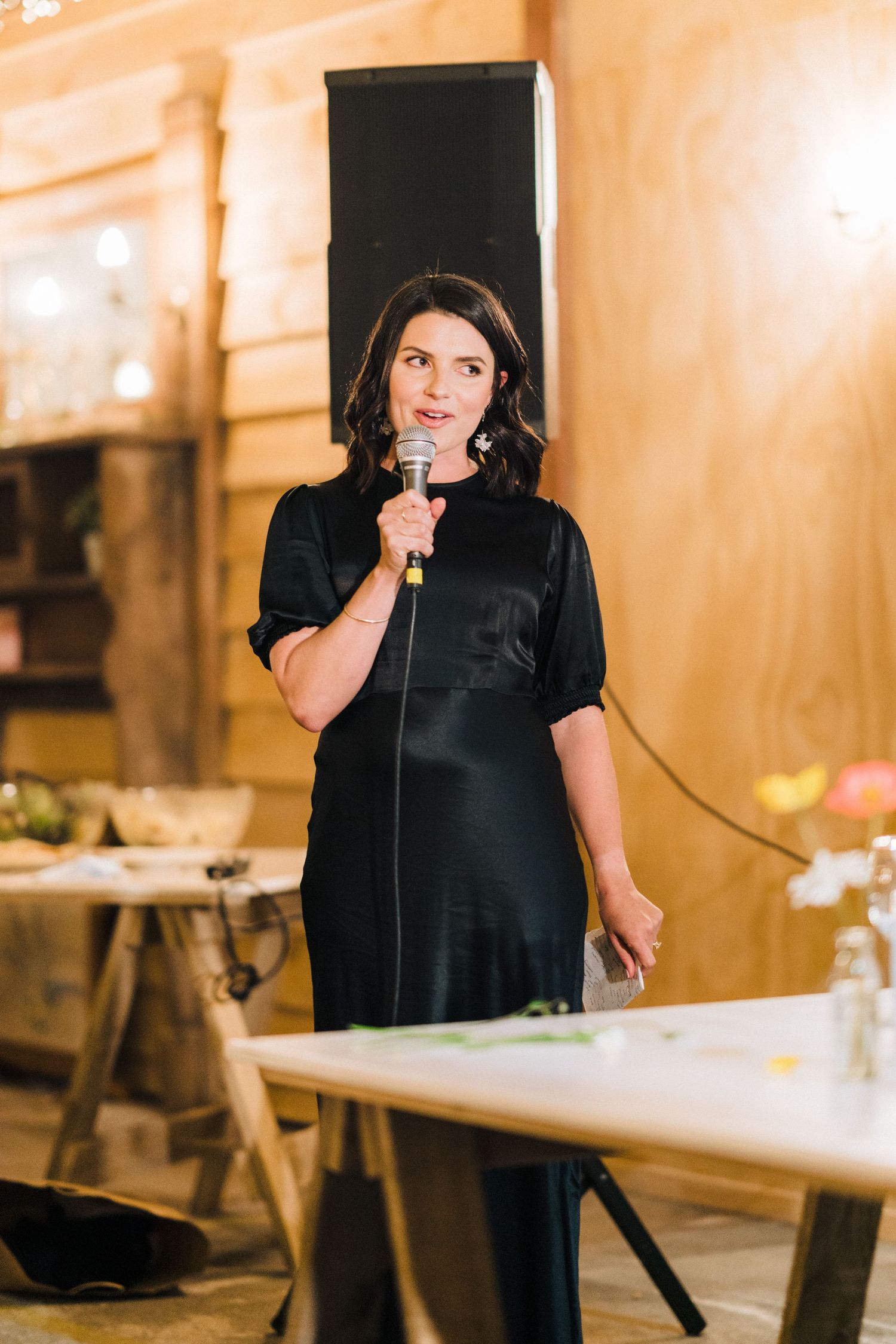 Speaker stands at podium in elegant black dress during an indoor event with warm lighting and rustic decor.