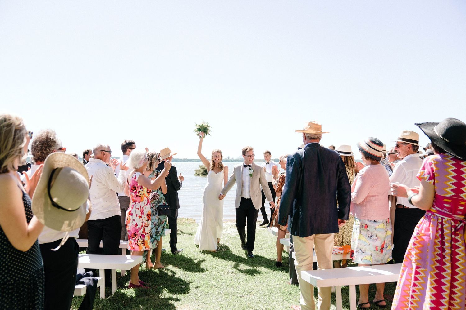 A bright sunny outdoor wedding celebration with guests standing on a grassy area near the ocean.