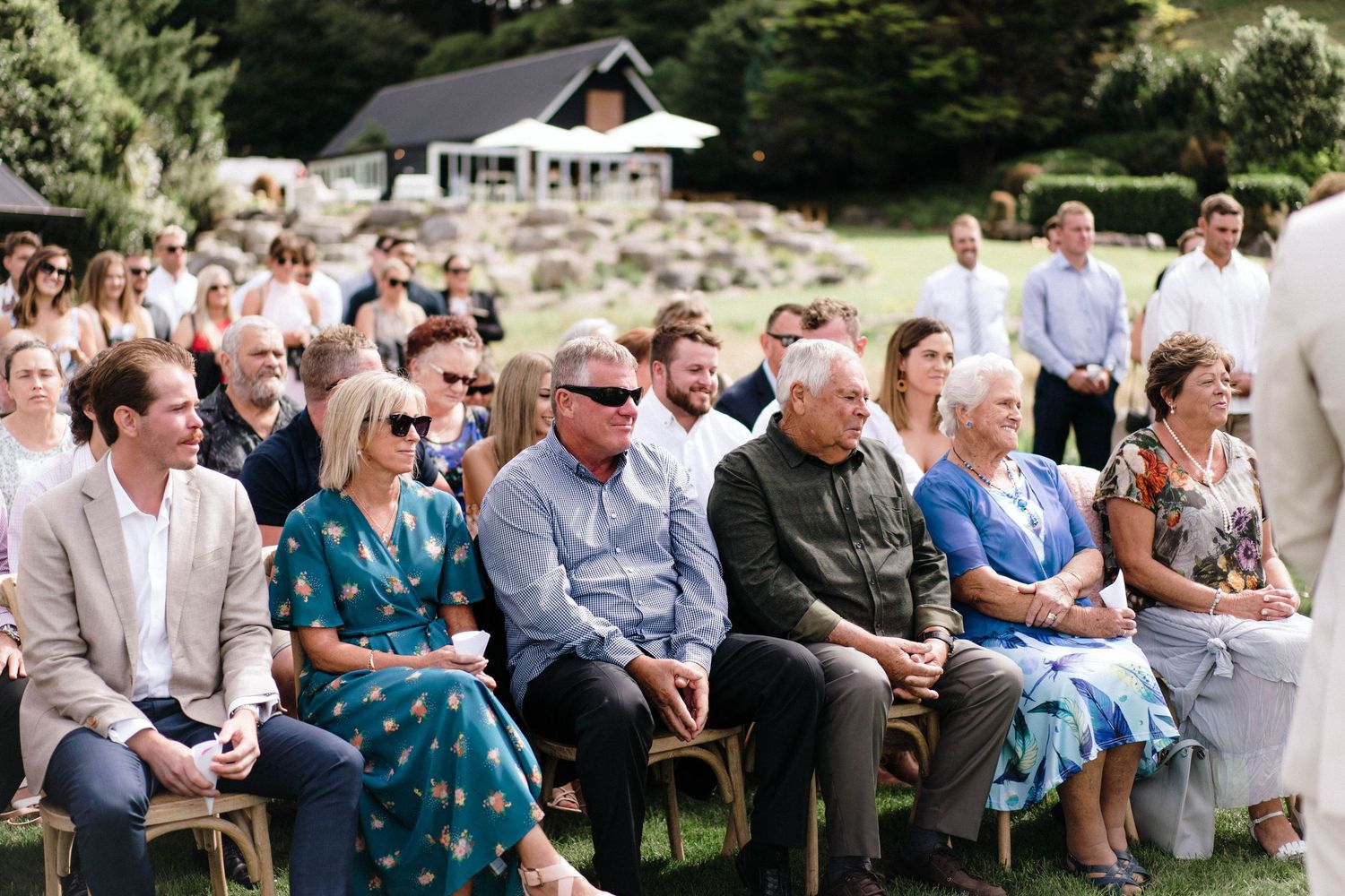Wedding guests seated outdoors on white chairs watching ceremony in natural setting.