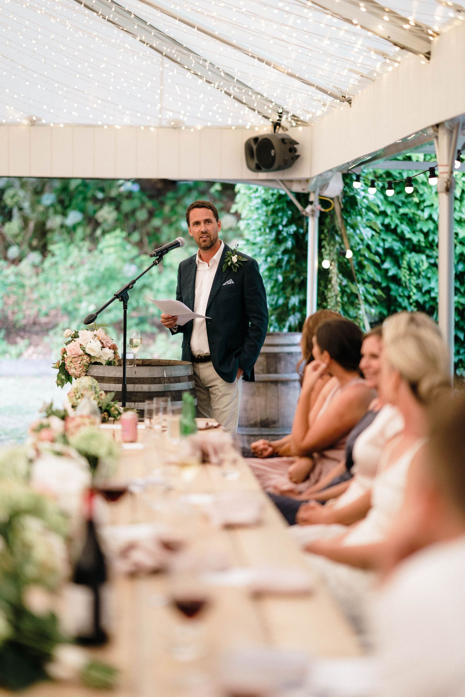 Wedding reception guests listen to speeches under a white tent outdoors.