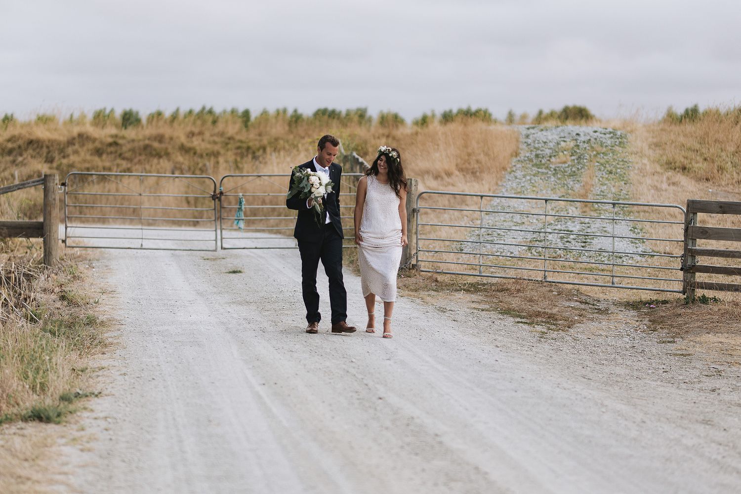 People silhouetted against setting sun walk on dusty country road with rustic wooden fences bordering the path.