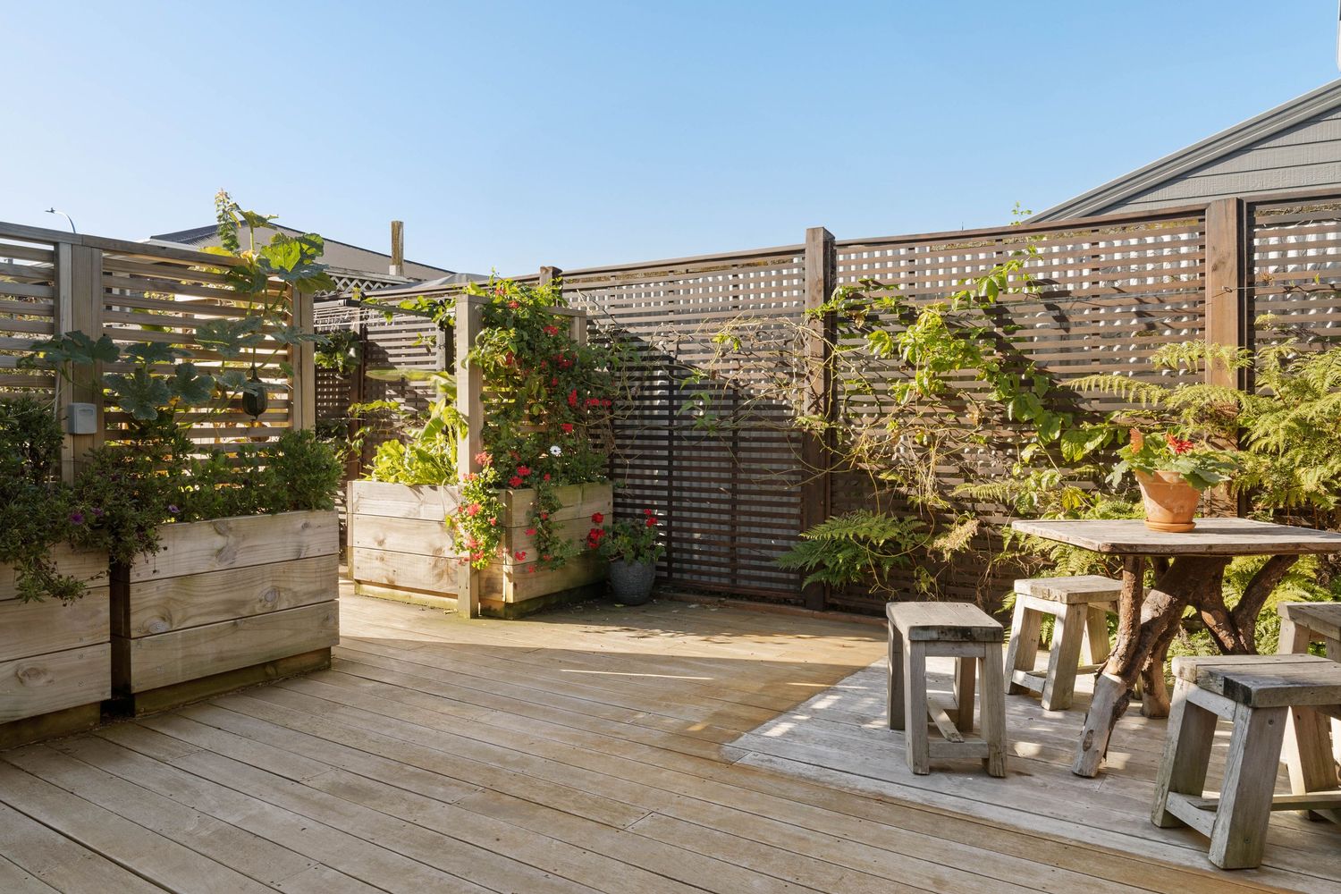 A modern rooftop deck features wooden flooring, lattice privacy screens, and potted plants under bright blue sky.