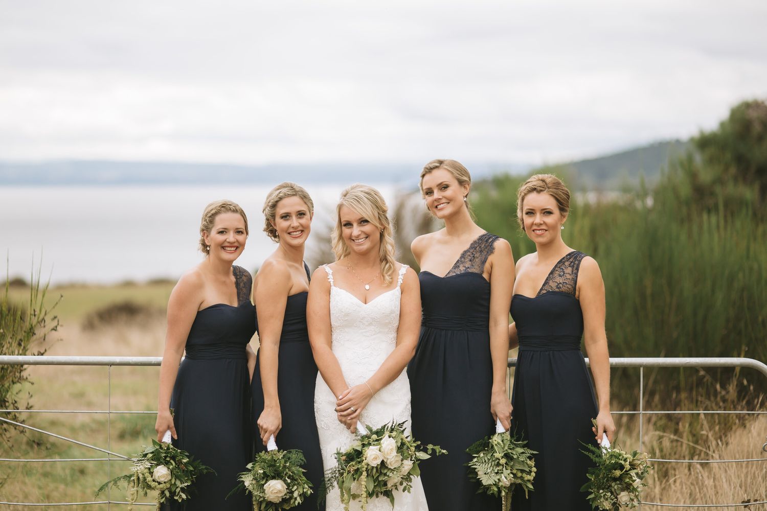 Bridesmaids in elegant black dresses pose with the bride holding white bouquets against a coastal backdrop.