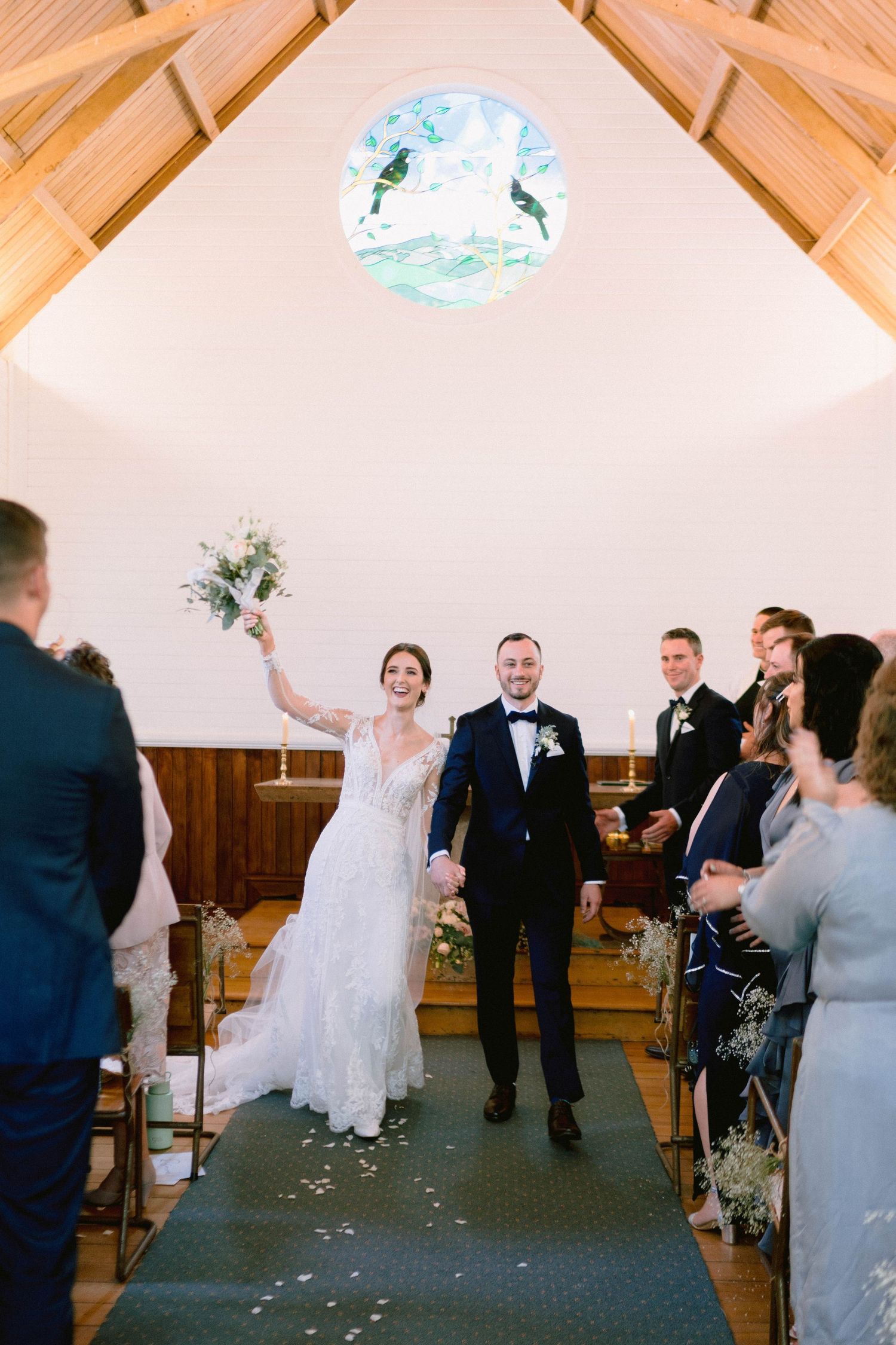 A newlywed couple celebrates at the altar in a rustic chapel with wooden beams and a round window.