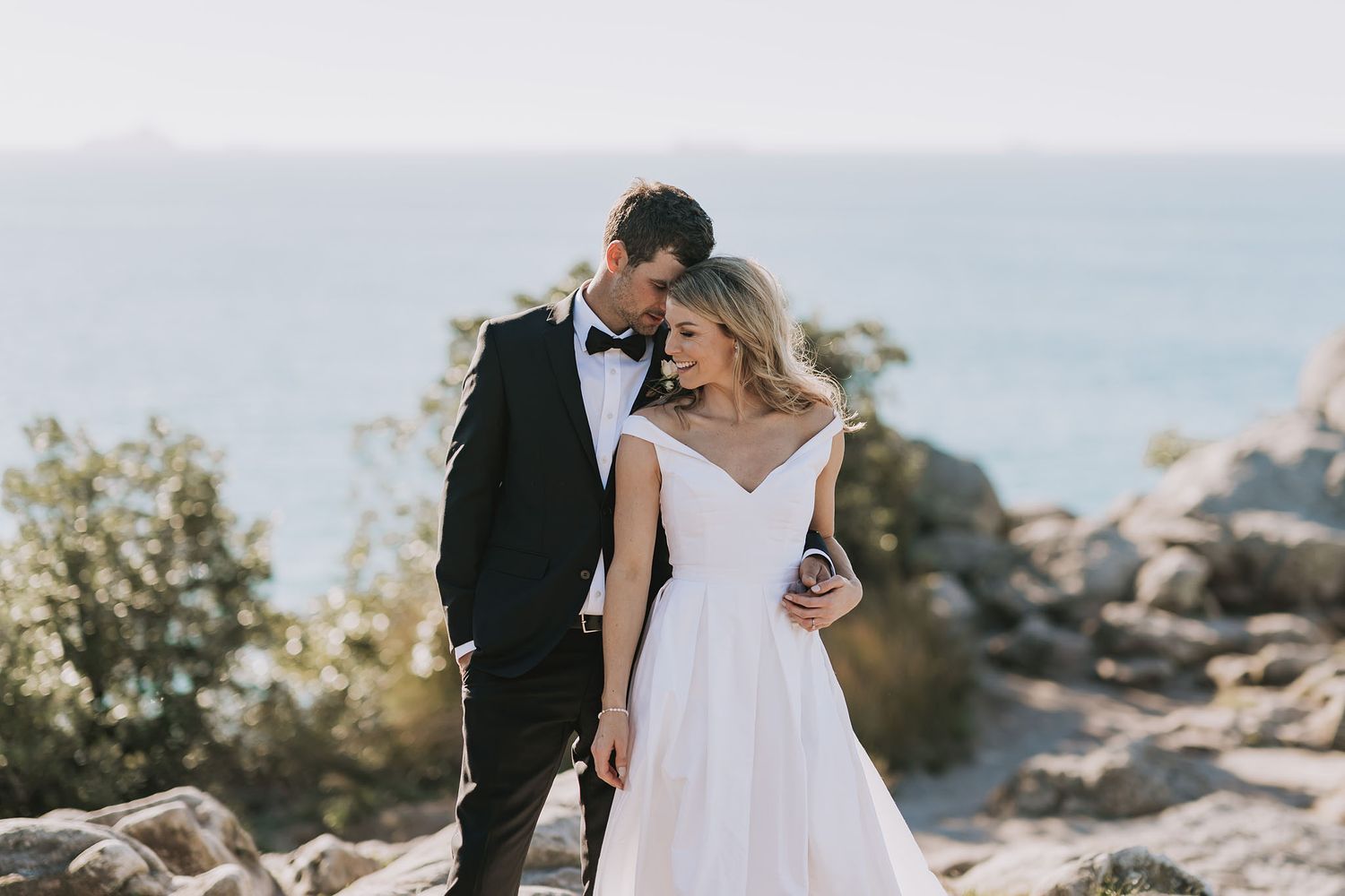 A romantic wedding couple embraces on rocky coastal cliffs overlooking the ocean at sunset.