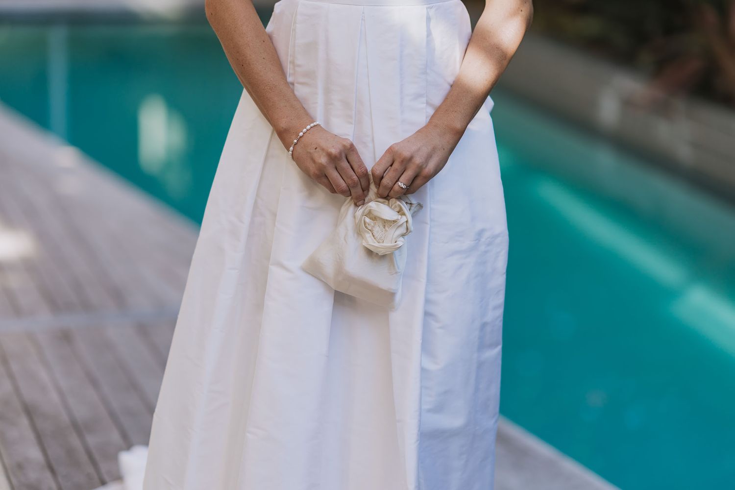Person in flowing white dress holds small purse while standing on wooden deck beside turquoise swimming pool.