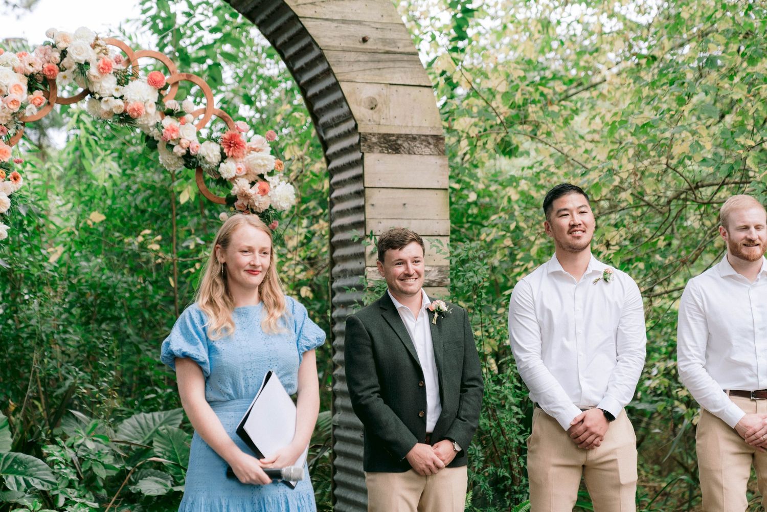 Wedding party stands under floral archway in garden setting wearing coordinated attire.