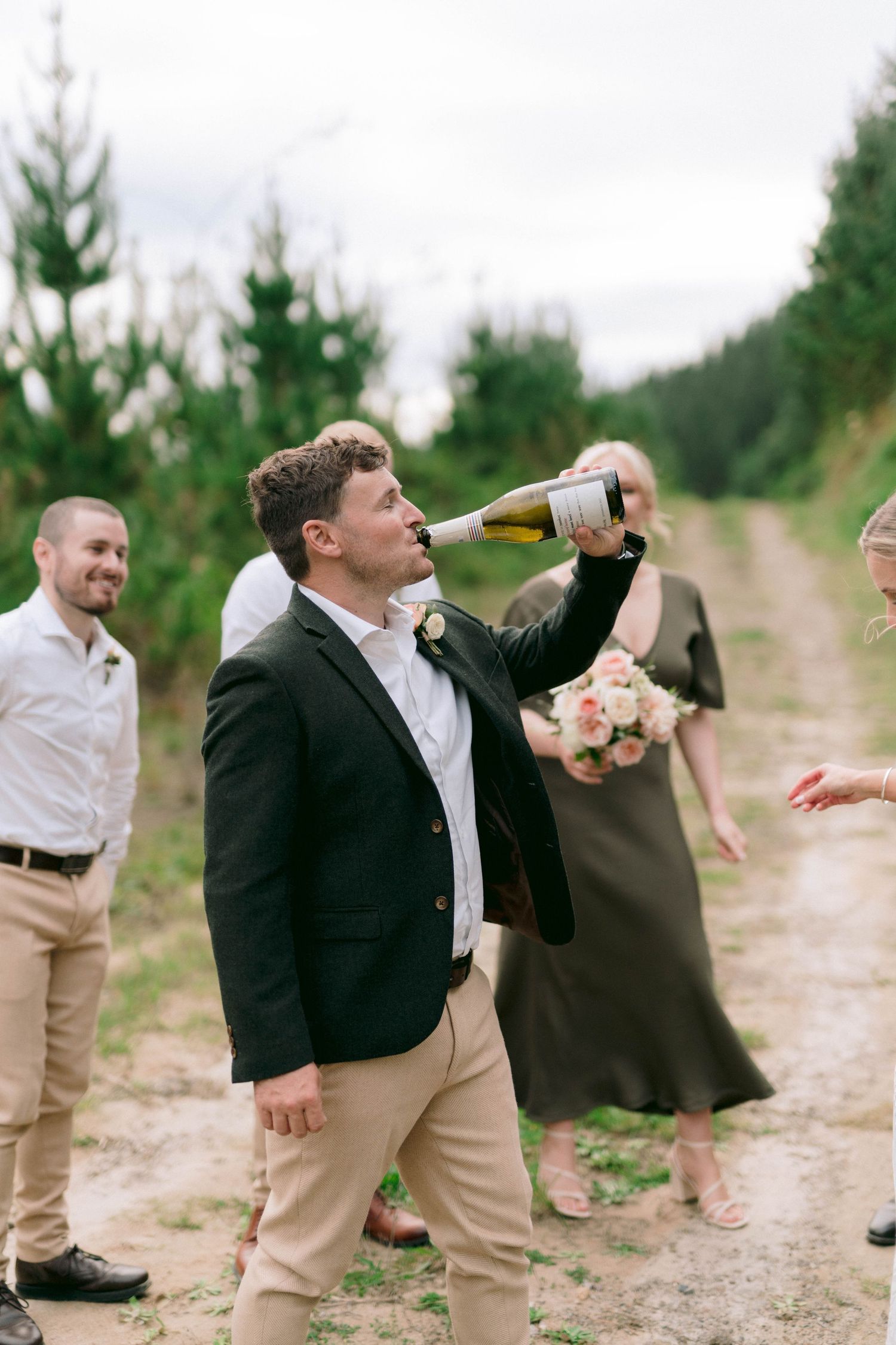 A wedding guest taking a drink from a bottle on a rustic dirt path surrounded by pine trees.