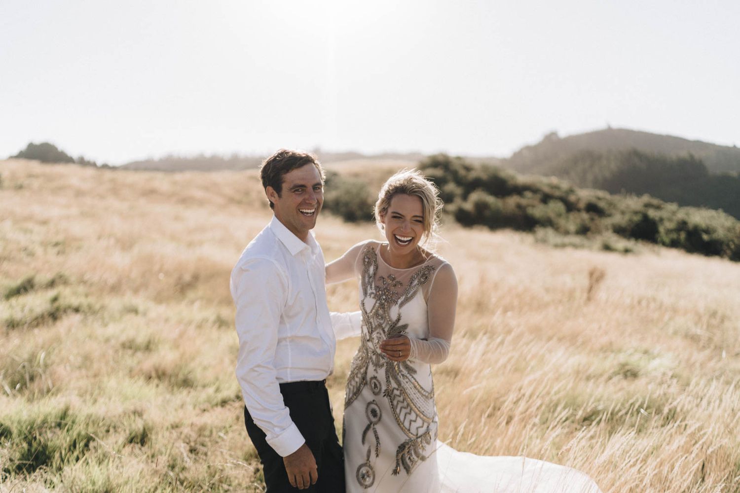 A romantic couple embraces in a golden sunlit field with rolling hills in the background during a photoshoot.