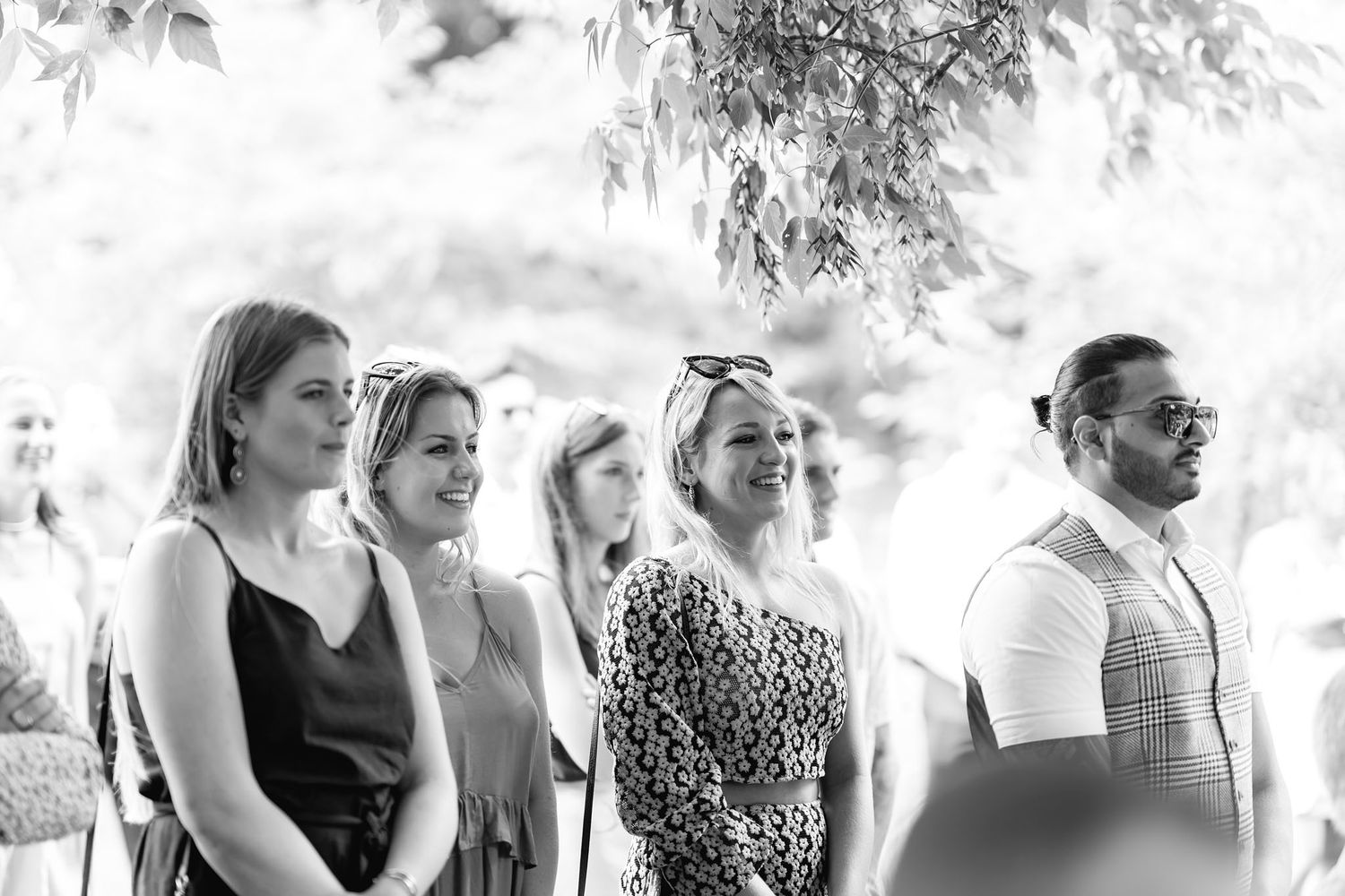 A black and white photo shows a group of formally dressed guests attending an outdoor event.