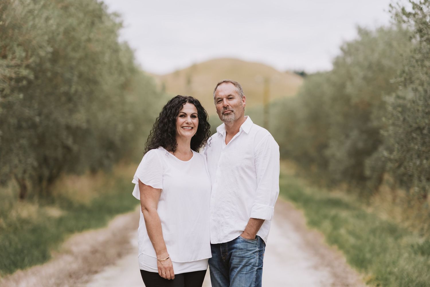 A couple in white casual clothes pose together on a peaceful dirt path lined with olive trees.