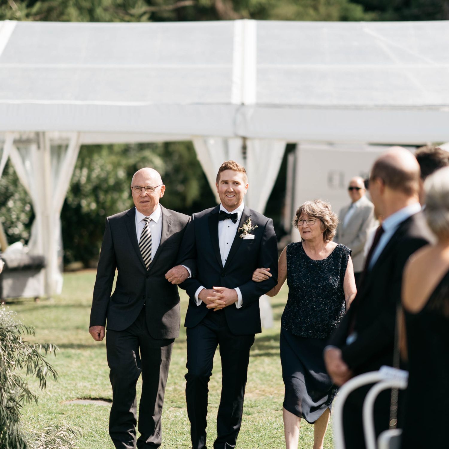A formal outdoor wedding party makes their way down the aisle under a white tent on a sunny day.