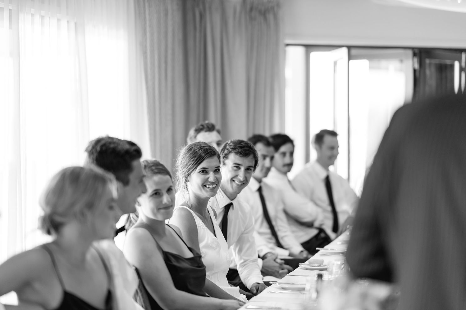 Wedding party members seated at a long table smile during a reception in black and white photography.