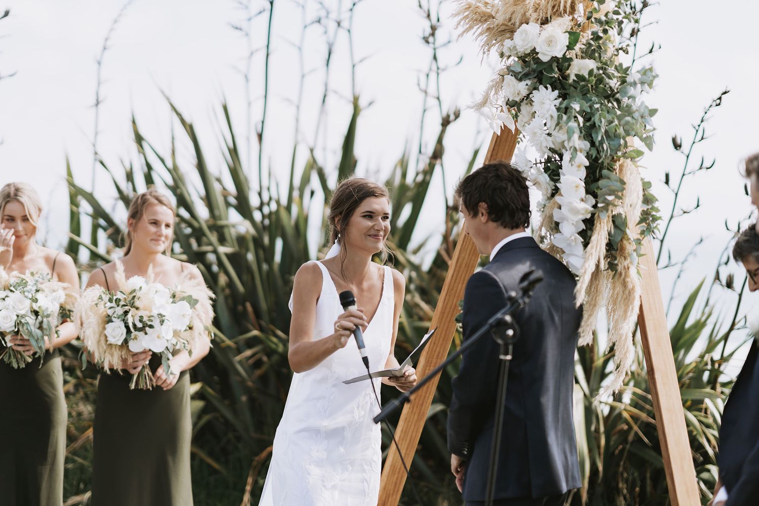 Couple exchanges vows at outdoor wedding ceremony surrounded by pampas grass and floral arch decorations.