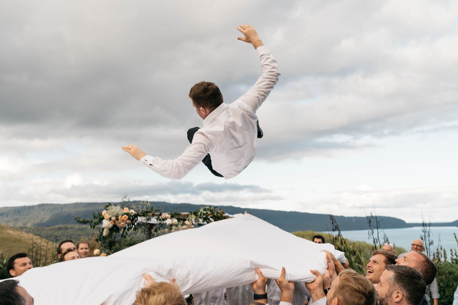 A person in white clothing leaps joyfully with arms raised against a scenic mountain landscape and cloudy sky backdrop.