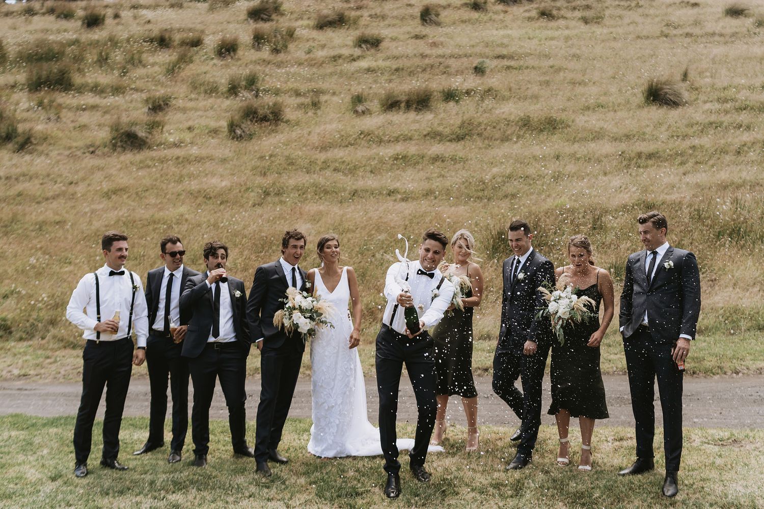 Wedding party stands in line along scenic hillside with warm brown grass as backdrop, showing formal and rustic attire.