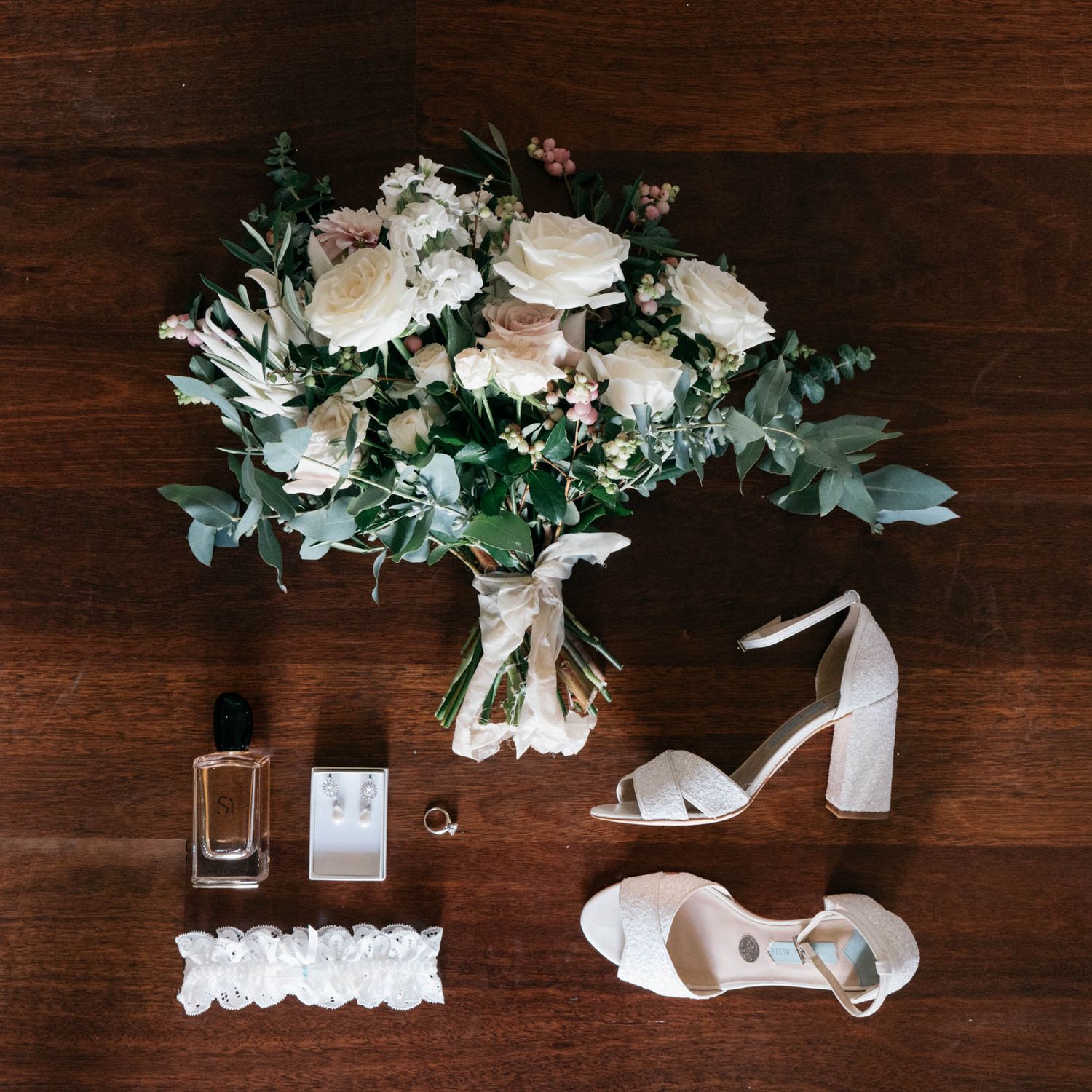 Wedding details with white floral arrangements, rings, perfume and ribbons laid out on dark wooden surface.