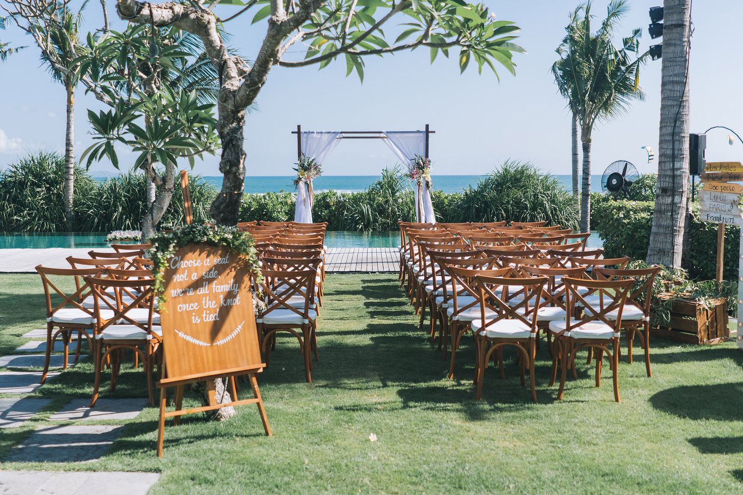 Outdoor wedding ceremony setup with wooden cross-back chairs and altar arch overlooking the ocean.