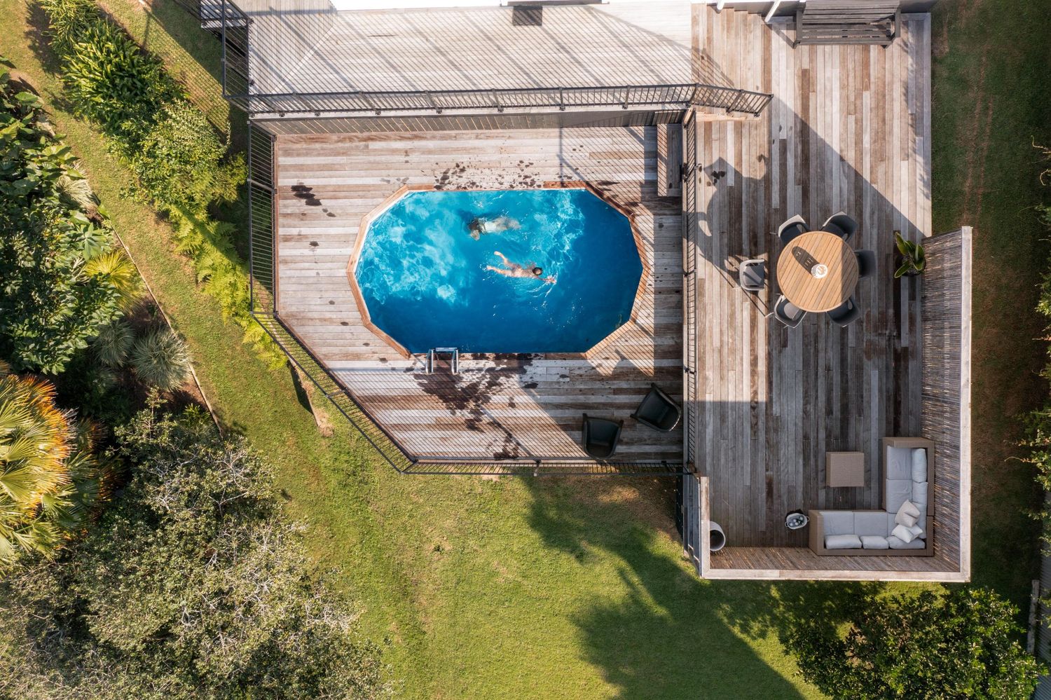 Aerial view of a bright blue swimming pool next to a wooden deck and grassy lawn in a backyard setting.