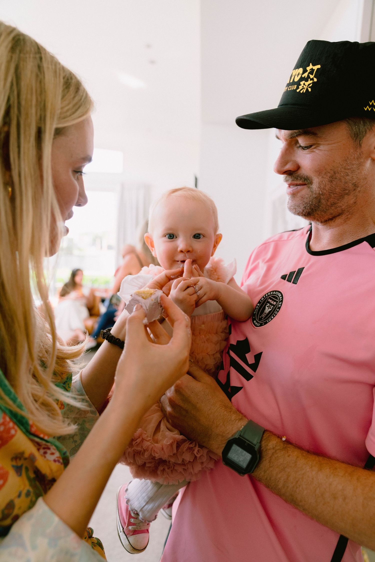 Baby in a pink dress being held while interacting with others at a casual gathering.