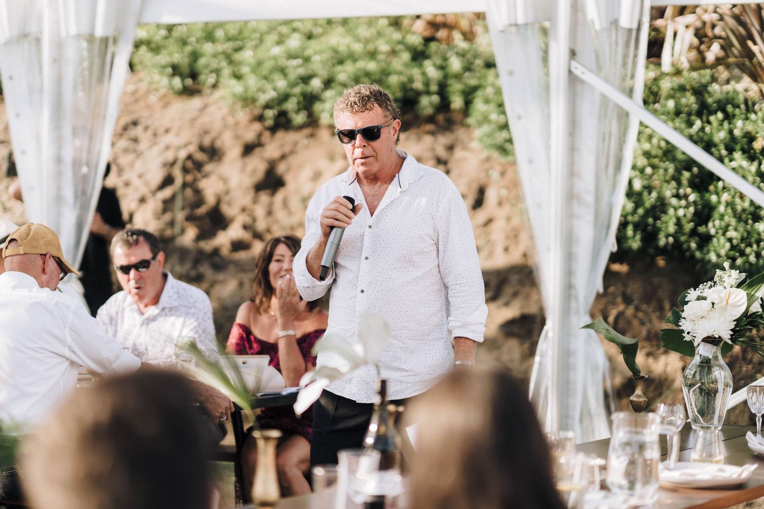 Wedding guests gather outdoors under white fabric draping at an elegant daytime celebration.