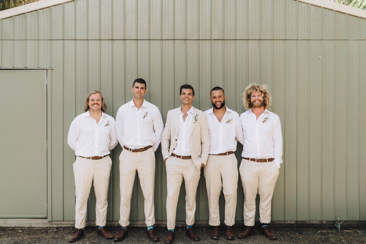 Groomsmen dressed in coordinating white and beige outfits pose against a gray building.