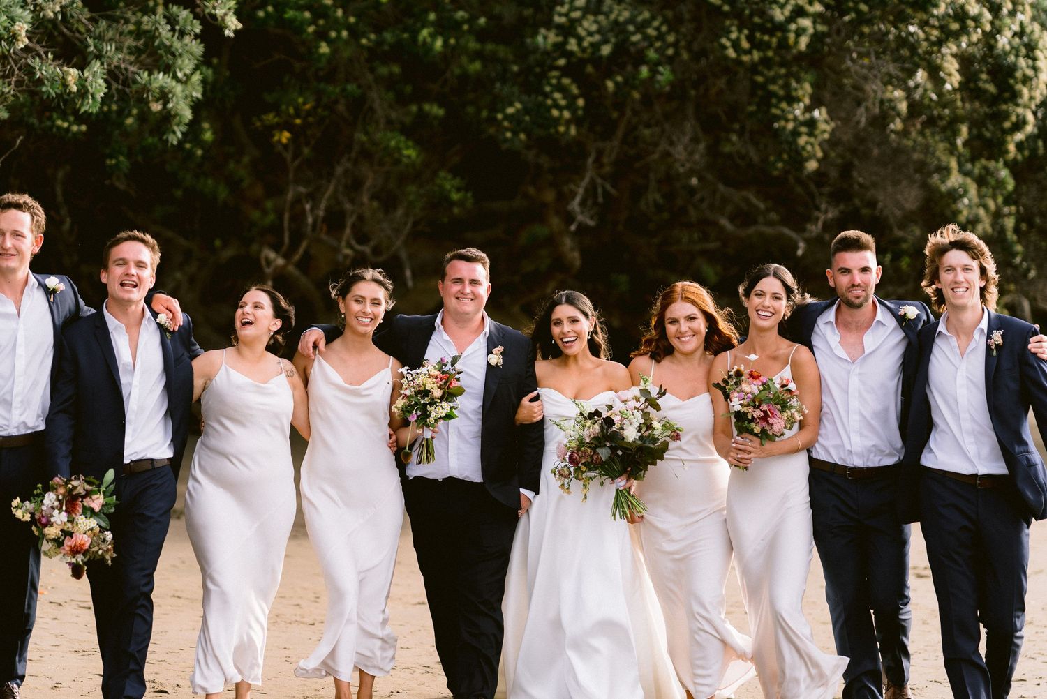 Wedding party in white dresses and dark suits poses together for formal group photo.