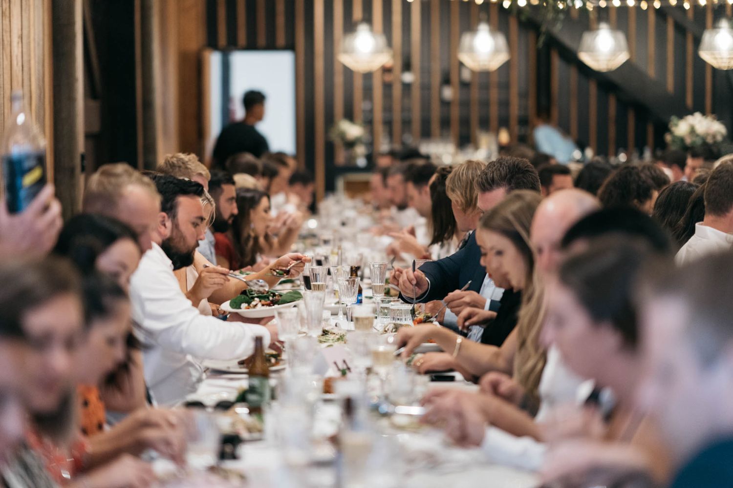 Diners gathered at a long communal table in a warmly lit restaurant enjoying a shared meal together.