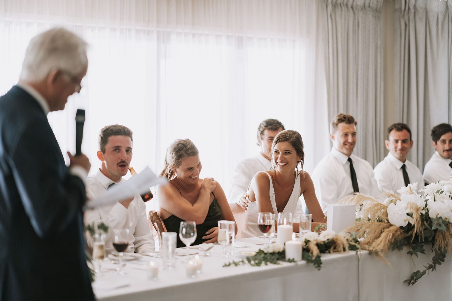 Wedding party members seated at head table laugh during speech at reception dinner.