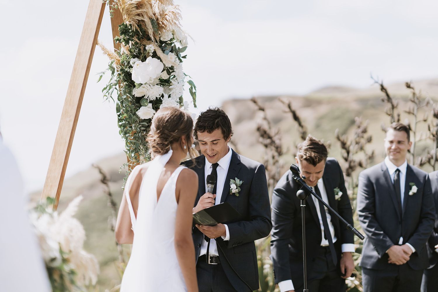 Elegant outdoor wedding ceremony taking place on a hilltop with white floral arch decorations and mountains in background.