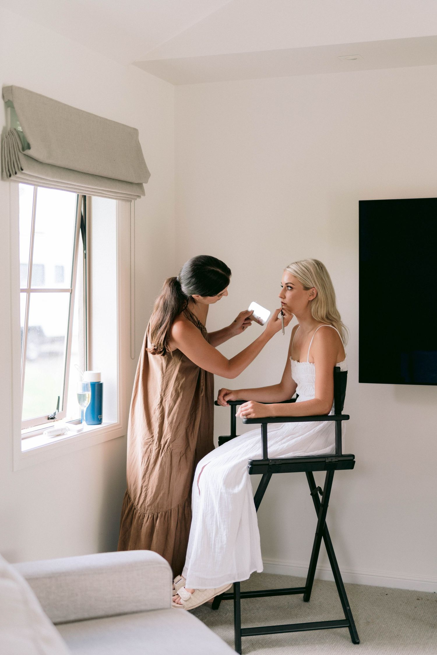 A makeup artist applies cosmetics to someone wearing a white gown seated in a director's chair by a window.