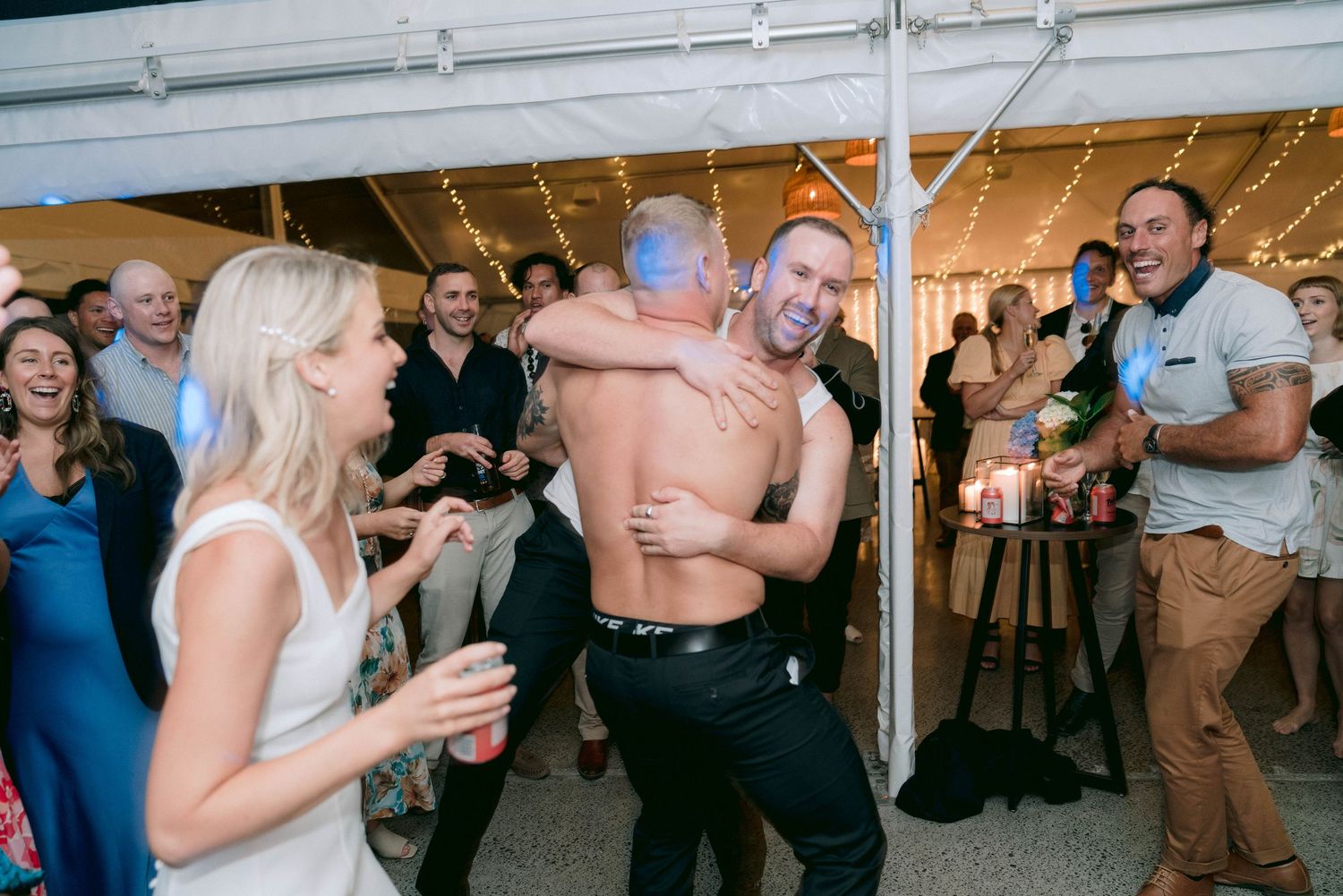 Wedding party and guests dancing together on a brightly lit dance floor under a white tent.