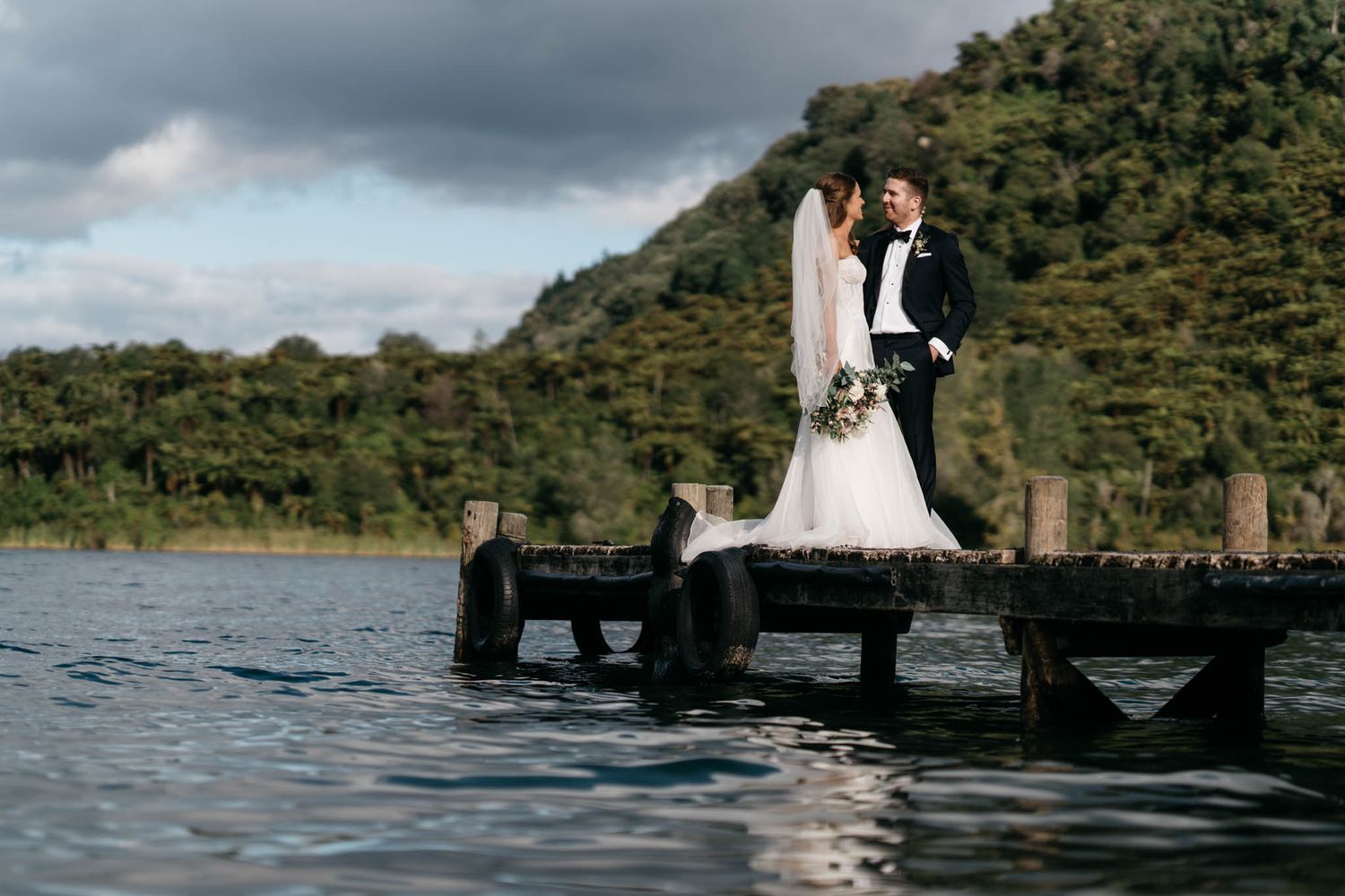 A couple in wedding attire stands together on a wooden dock by a serene lake with green hills in the background.