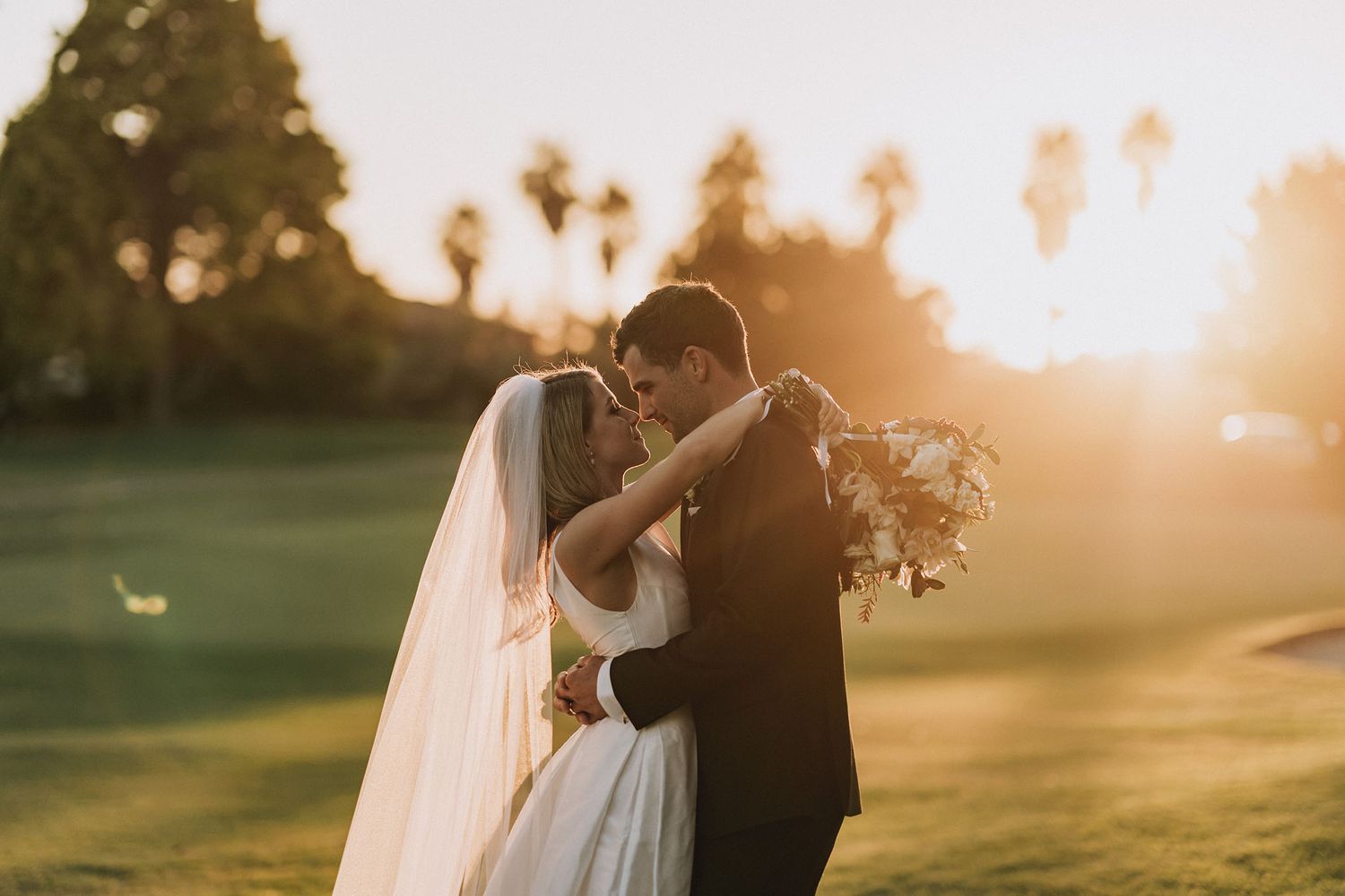 A couple shares a romantic kiss at sunset on their wedding day on a grassy field with palm trees in the background.