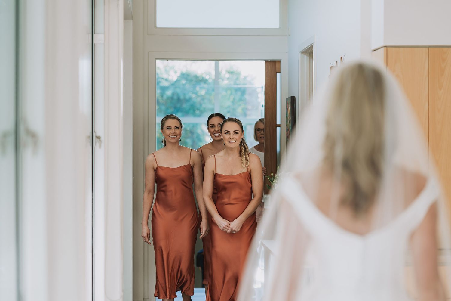 Bridesmaids in burnt orange dresses line a bright hallway as they watch someone in white at the end.