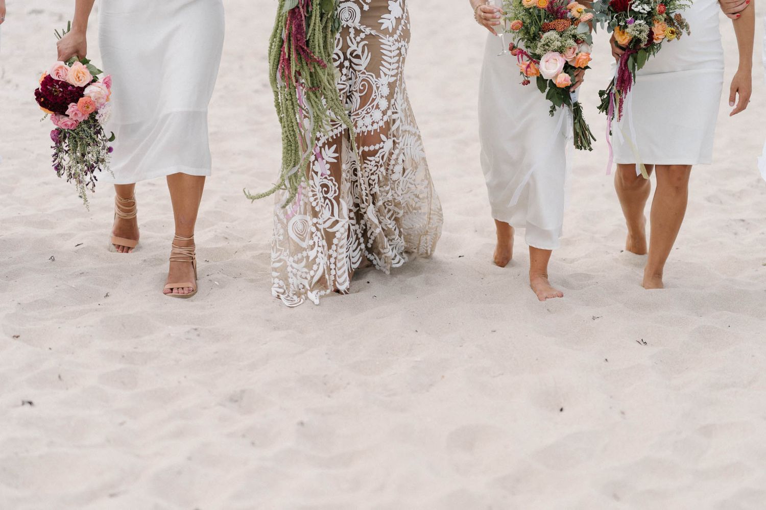 Wedding party wearing white dresses and holding pink floral bouquets walks along sandy beach during sunset celebration.