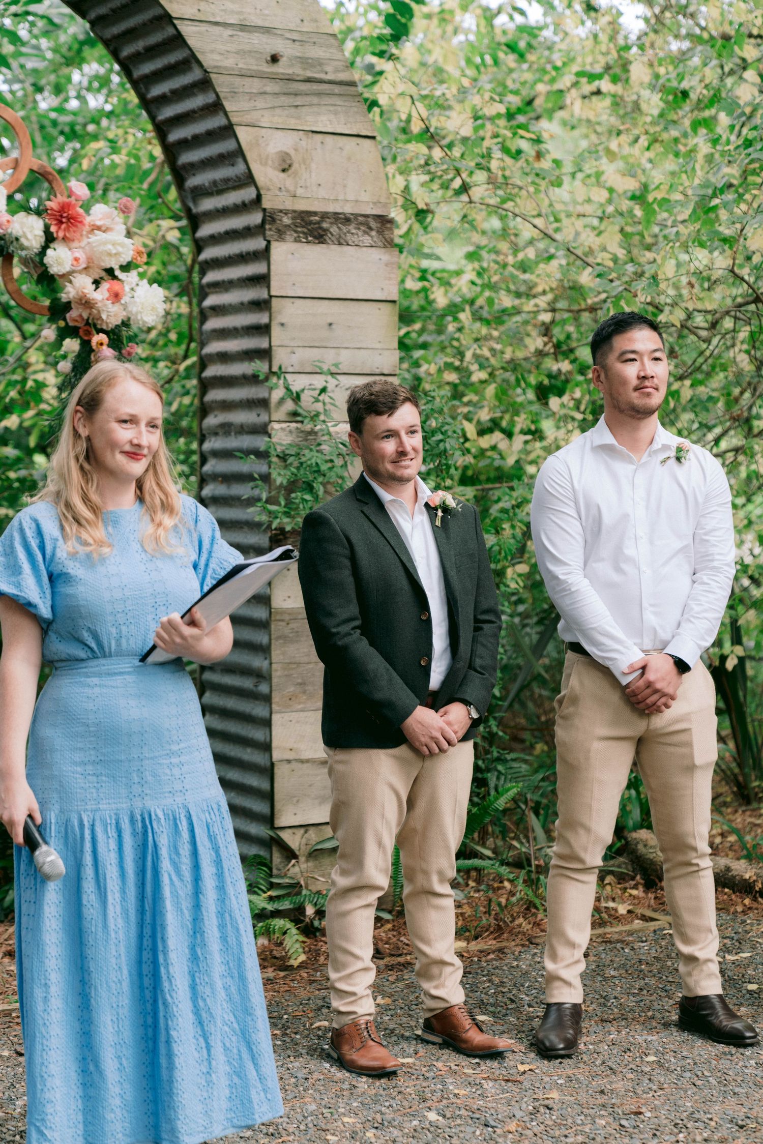 Three attendants stand in front of a rustic garden arch with blooming flowers.