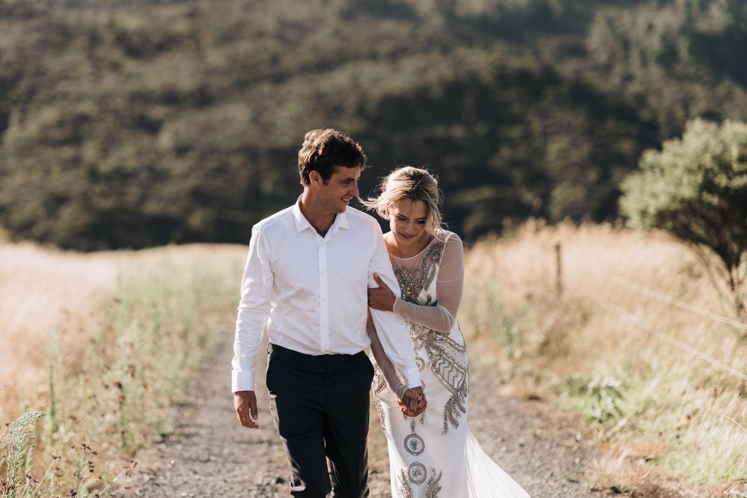 A couple walks hand in hand through a golden mountain meadow, with the bride wearing a white lace wedding gown.