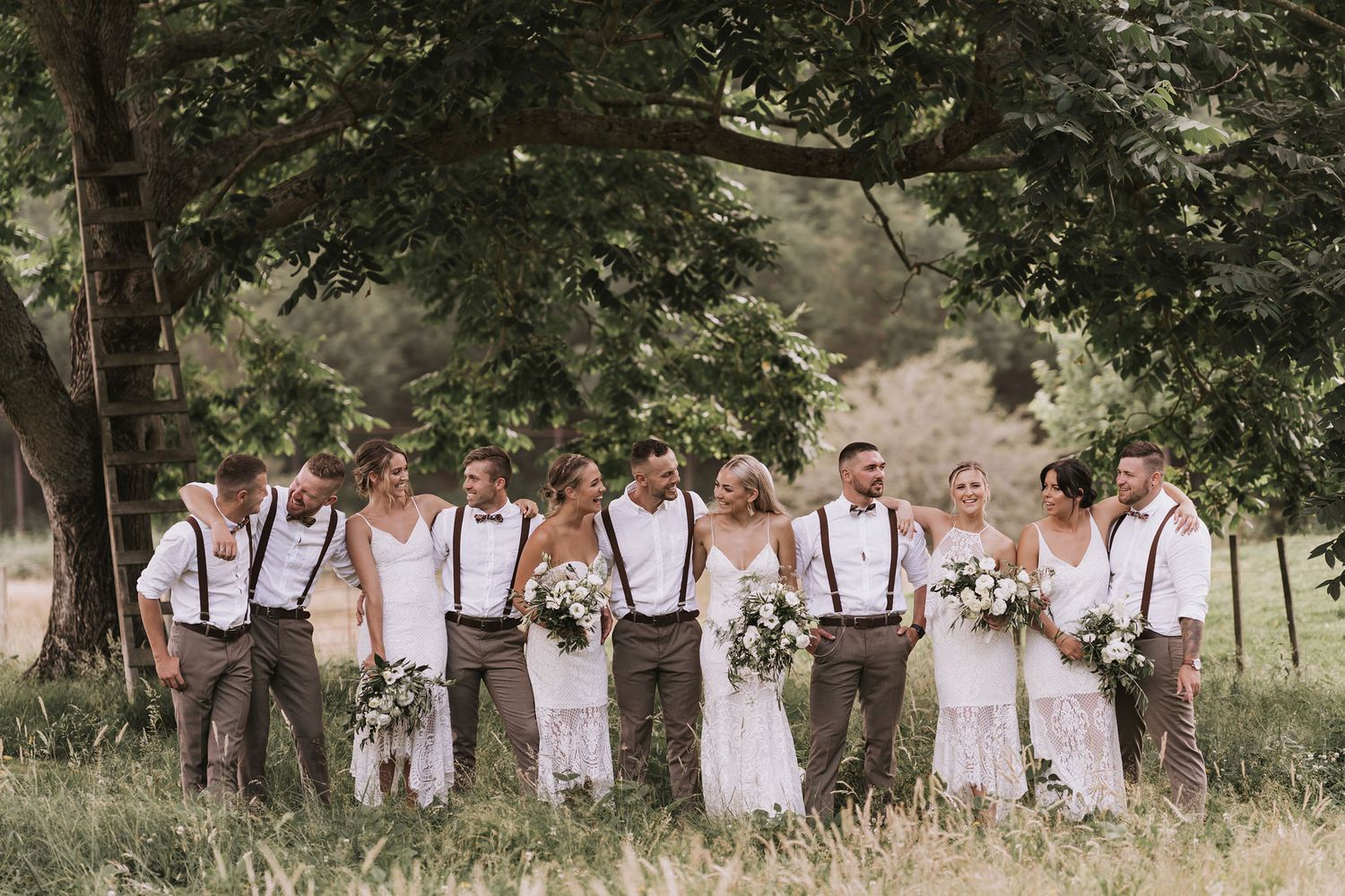 Wedding party in white dresses and suspenders posing together under trees in a natural outdoor setting.