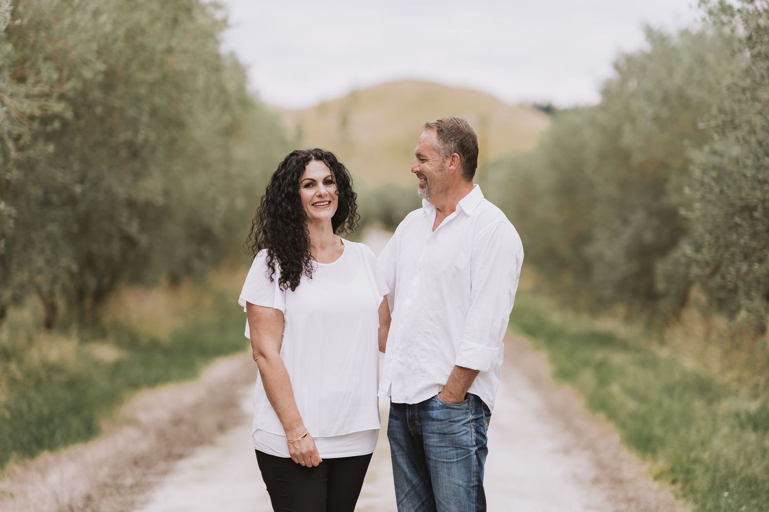 A loving couple walks hand in hand down a scenic dirt path surrounded by green foliage on a bright summer day.