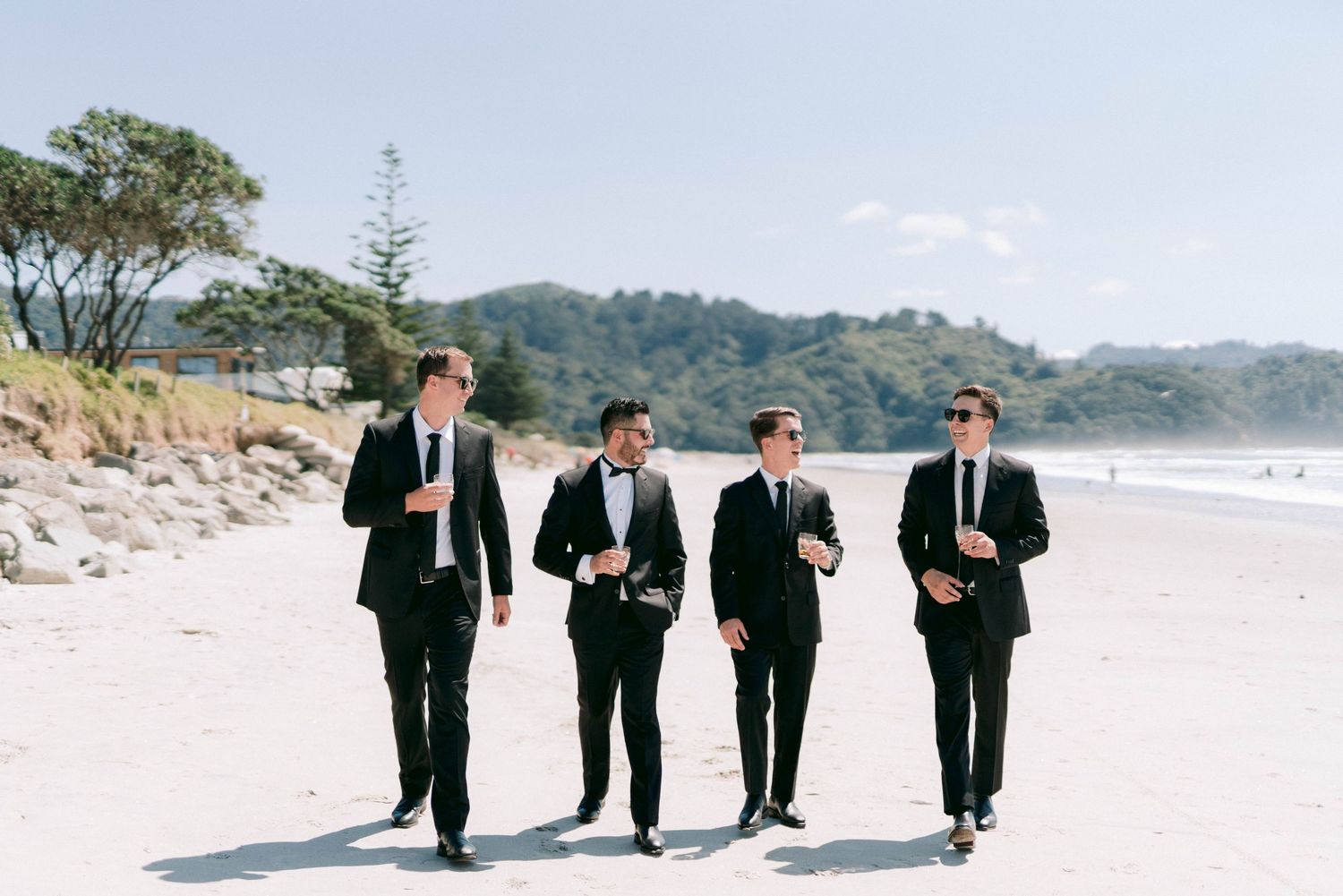 A group of four people in black suits walking along a scenic beach with mountains in the background.