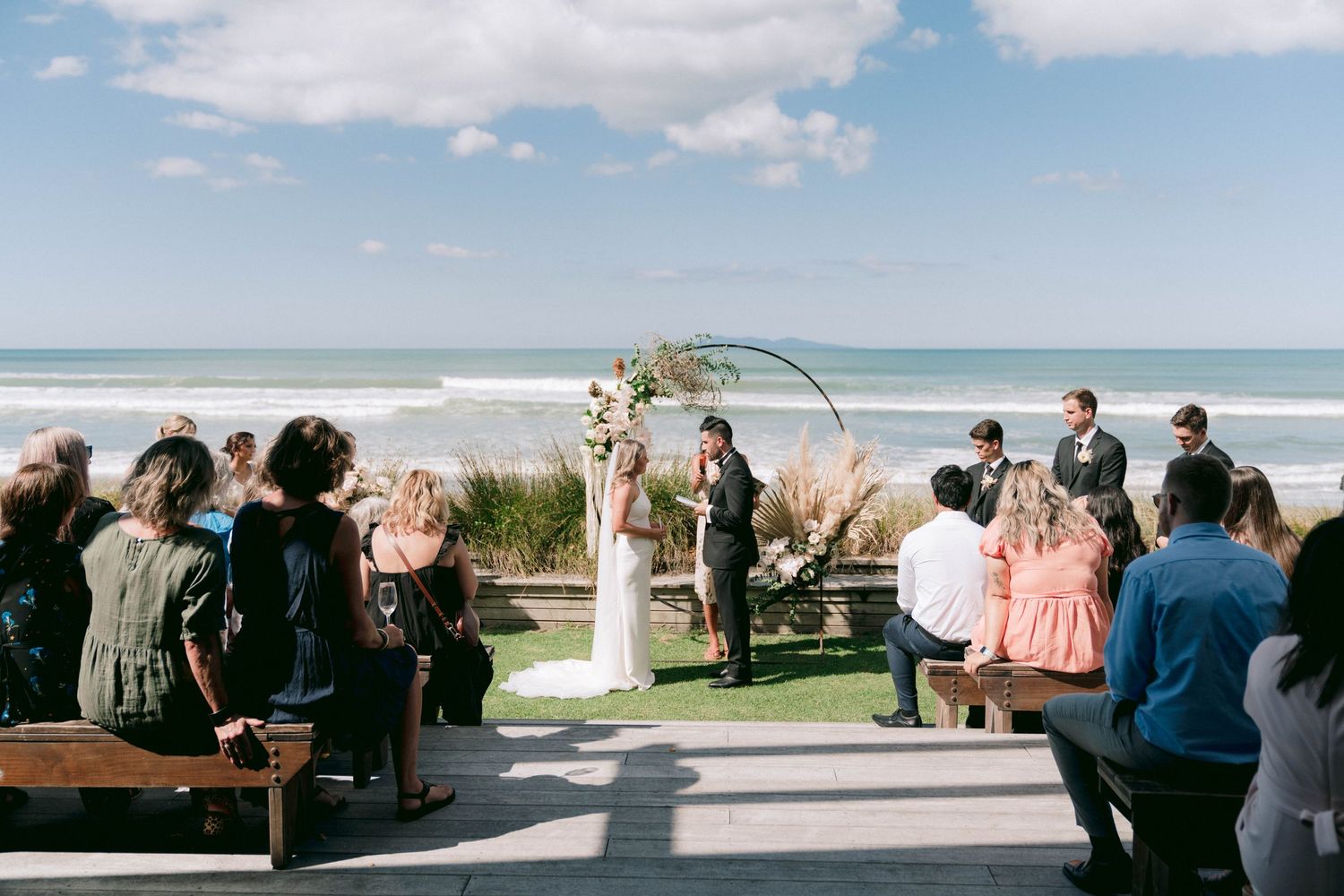 Oceanfront wedding ceremony with white arch and guests seated on wooden benches.