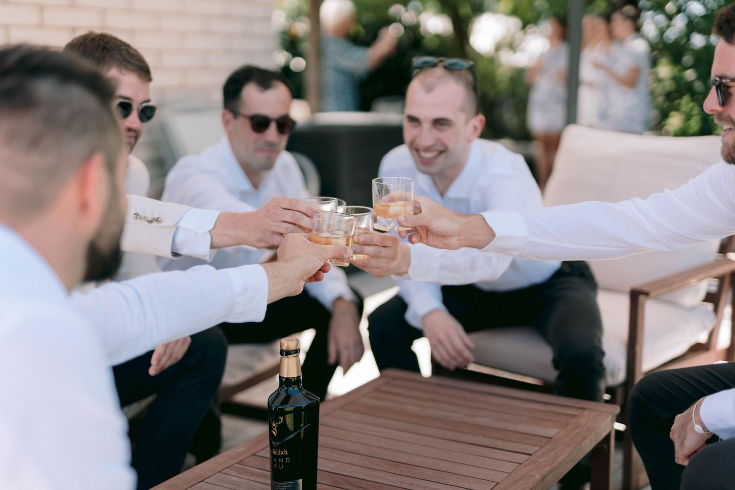 A group of friends in white attire toast their drinks together at an outdoor gathering.