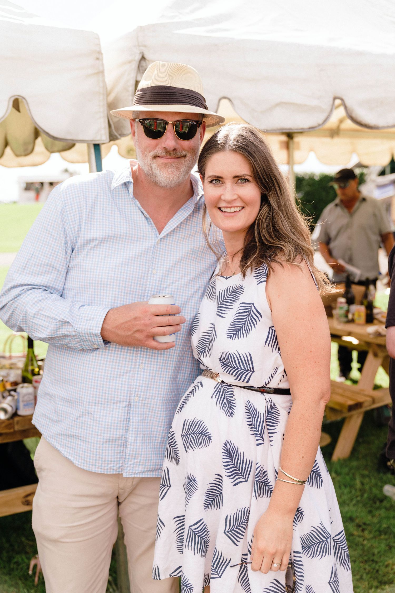 A couple in summer attire poses together at an outdoor event under white tents.