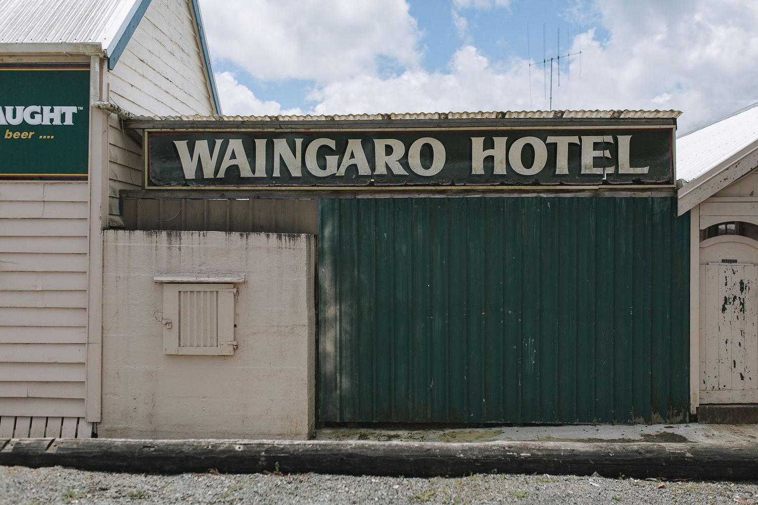 Rustic white wooden building with green signage for Waingaro Hotel against a blue sky with clouds.