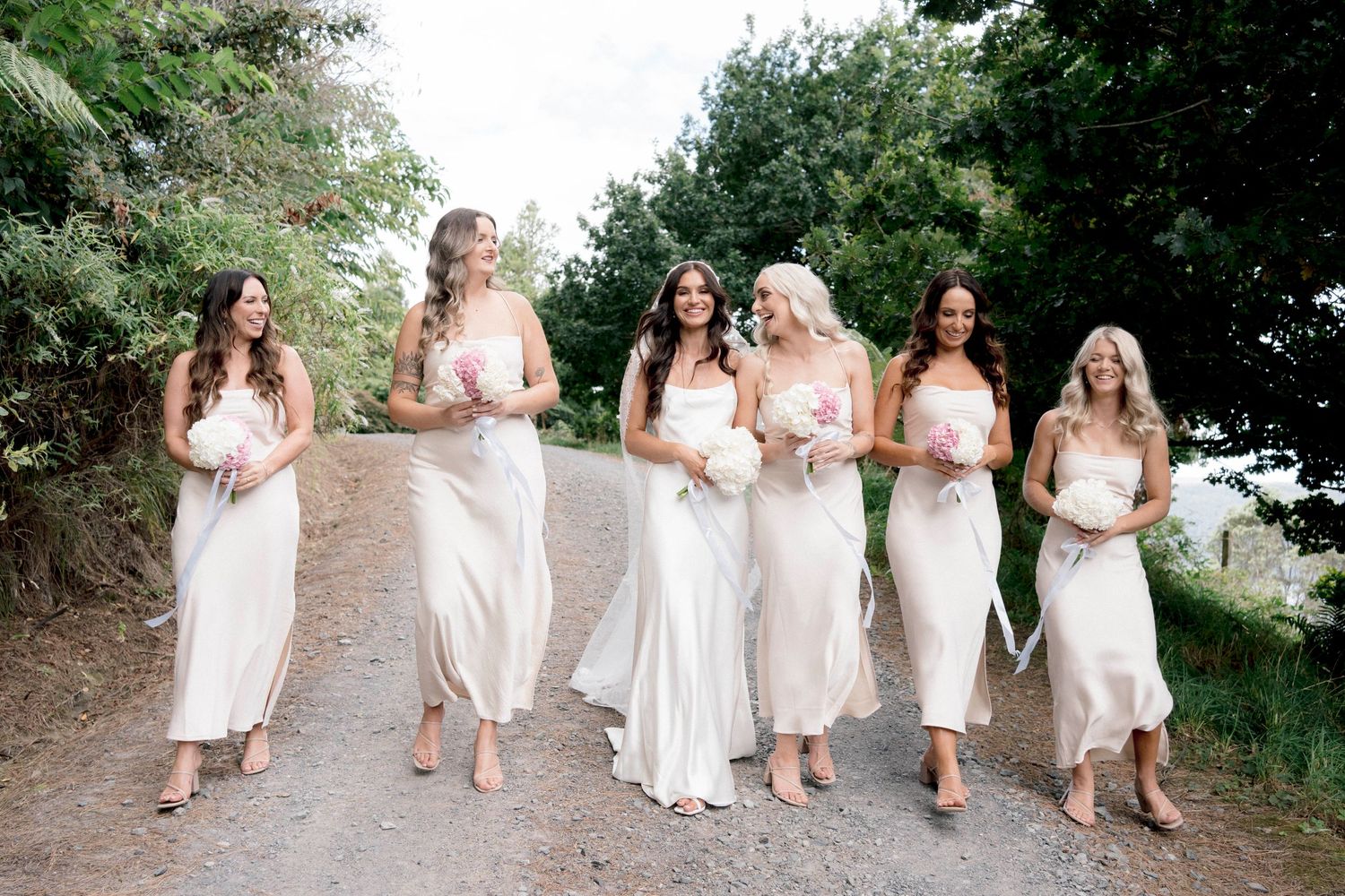 Bridesmaids in matching white dresses walk together on dirt path during wedding photoshoot.