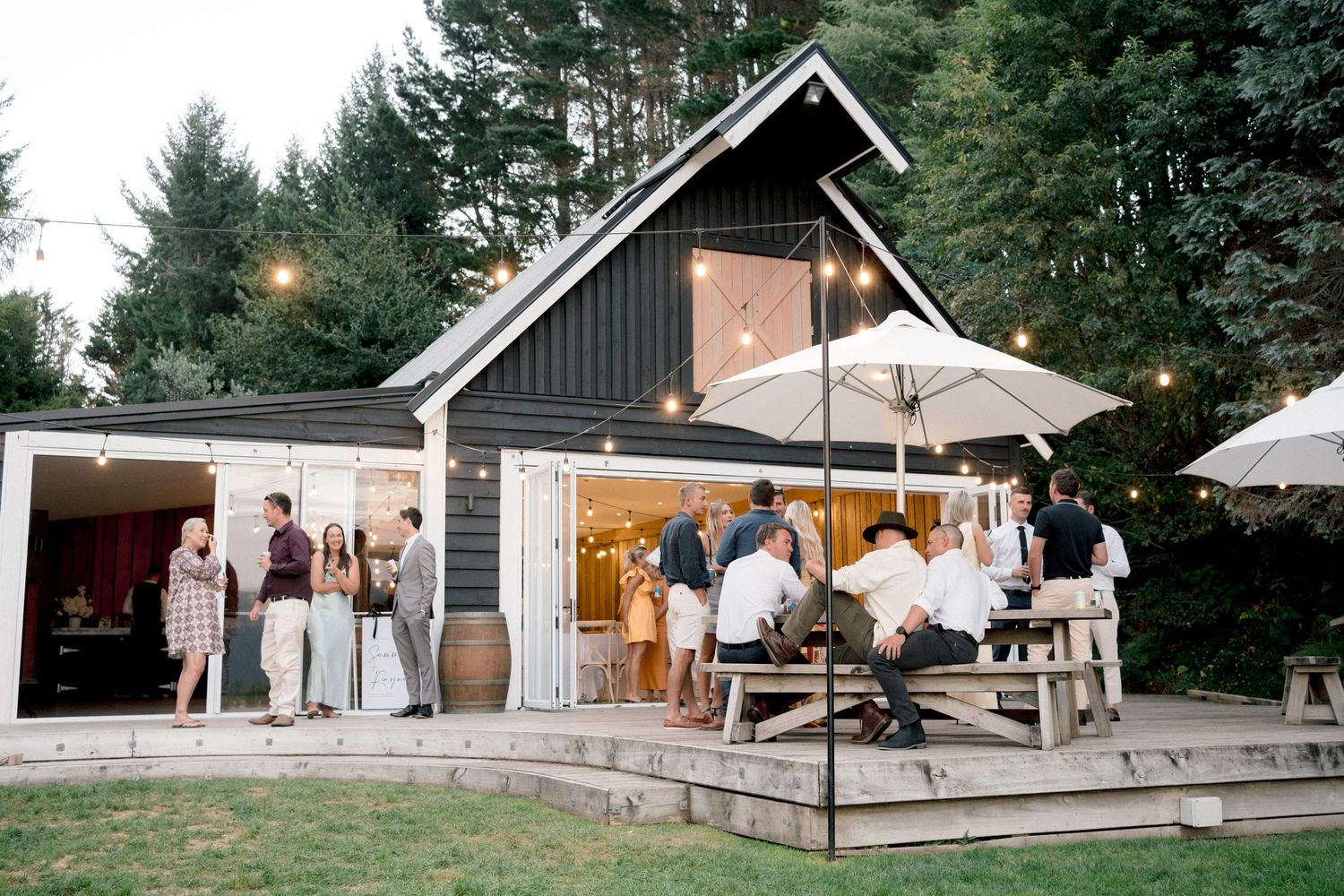 Guests gather outside a charming black barn venue with string lights and umbrellas at dusk.