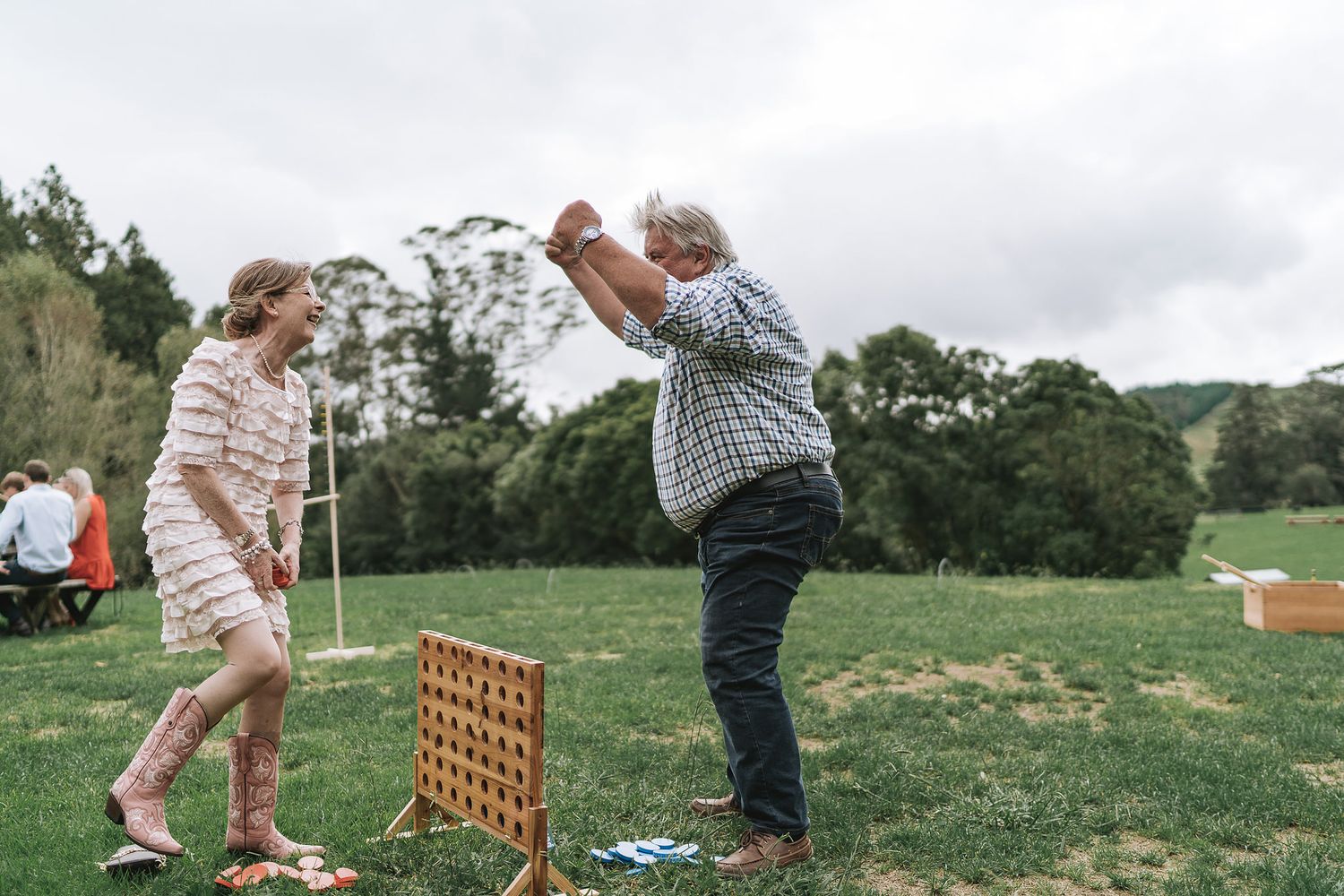 Two people play a life-sized Connect Four game on grass with trees in the background during a cloudy day.