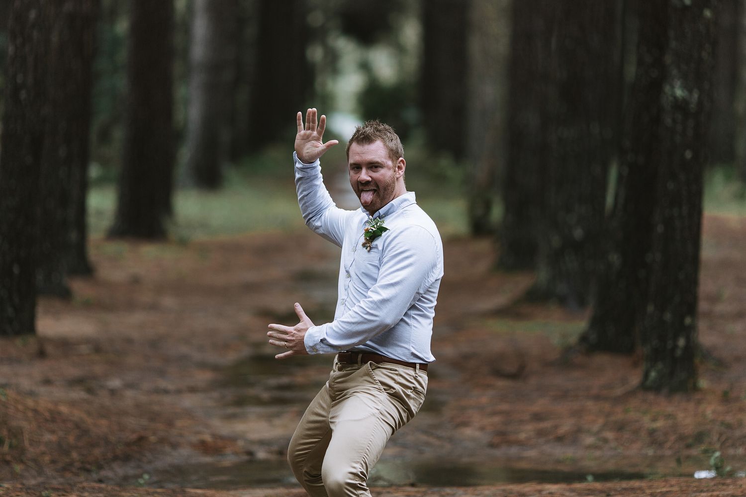 A joyful person wearing khaki pants and white dress shirt jumps with excitement in a pine forest setting.